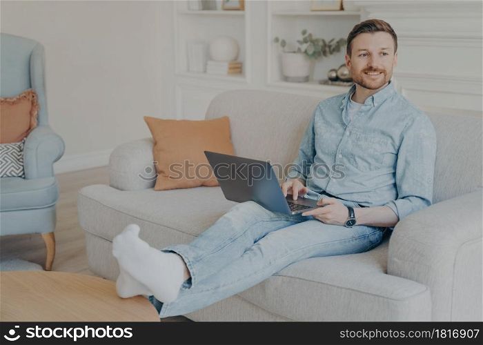 Young man with stubble surfing web on his laptop while sitting on couch, resting legs on coffee table, relaxing after hard work session on project, taking break from working. Relaxed young man surfing web while sitting on couch