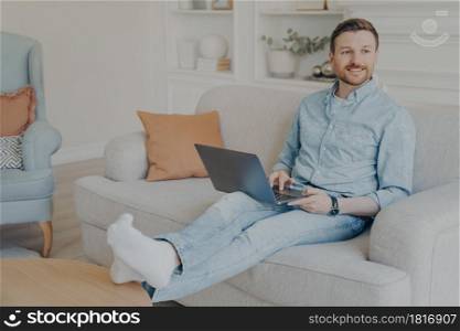 Young man with stubble surfing web on his laptop while sitting on couch, resting legs on coffee table, relaxing after hard work session on project, taking break from working. Relaxed young man surfing web while sitting on couch