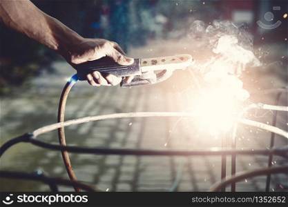 Young man worker welding iron pieces at work with protective goggles. Building construction concept.
