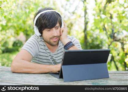 young man working with a tablet pc listening music with headphones on a wooden table, outdoor