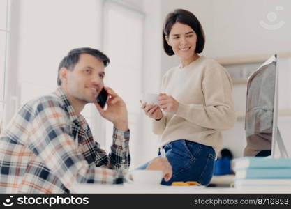 Young married couple study contract for new house, check documents for buying property, man discusses something with partner via cellphone, happy woman drinks tea and looks in computer monitor