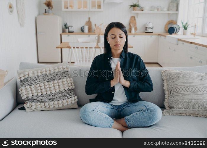 Young mixed race woman is practicing yoga at home. Girl sitting on couch in lotus pose and meditating with her eyes closed. Tranquility and stress relief. Peaceful mind, zen and wellness.. Young mixed race woman is practicing yoga, sitting on couch in lotus pose with her eyes closed.
