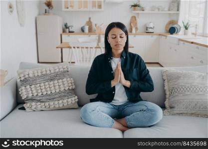 Young mixed race woman is practicing yoga at home. Girl sitting on couch in lotus pose and meditating with her eyes closed. Tranquility and stress relief. Peaceful mind, zen and wellness.. Young mixed race woman is practicing yoga, sitting on couch in lotus pose with her eyes closed.