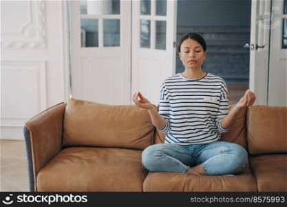 Young mixed race woman is practicing yoga at home. Lady is sitting on couch in lotus pose and meditating with her eyes closed. Stress relief and relaxation. Mental health and body care.. Young woman is practicing yoga at home. Lady is sitting on couch in lotus pose and meditating.