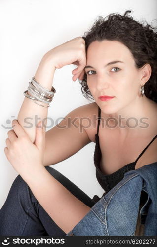 Young model in her mid twenties sitting on a chair with a blue denim jacket hanging over the back of the chair, head resting against hand, on white background.