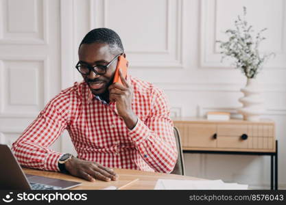 Young modern african businessman talking via mobile phone with client or partner while working remotely at home, pleased male afro american office worker making call using smartphone at work. Young modern african businessman talking via mobile phone with client while working remotely