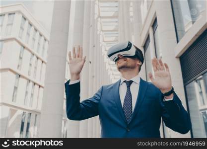 Young modern businessman in VR goggles on head working in augmented reality world and smiling, playing games in 3d goggles while standing outdoors, gesturing and interacting with cyberspace. Young modern businessman in VR goggles on head working in augmented reality world and smiling