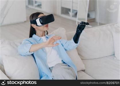 Young modern disabled girl with bionic prosthesis arm and virtual reality glasses playing video game sitting resting on sofa in living room at home. Disability and medical high tech concept.. Young disabled girl with bionic prosthesis arm, virtual reality glasses playing video game at home