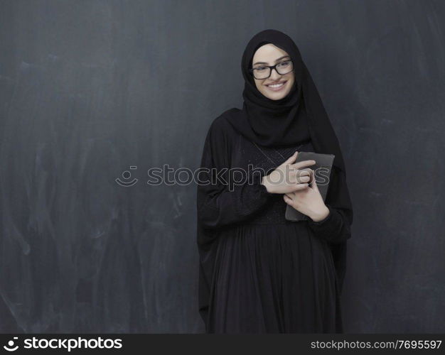 young modern muslim business woman using tablet computer wearing glasses and hijab clothes in front of black chalkboard