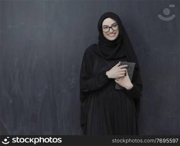 young modern muslim business woman using tablet computer wearing glasses and hijab clothes in front of black chalkboard