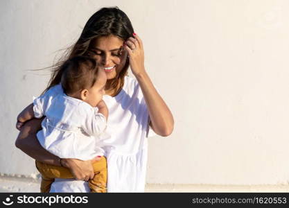 Young mom holding her little baby against white wall in sunny day