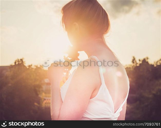 Young mom in a stylish, white dress standing on a background of clear sky and sunlight, holding a beautiful red rose flower in her hands. Concept of a happy family and motherhood. Young mom in a stylish, white dress