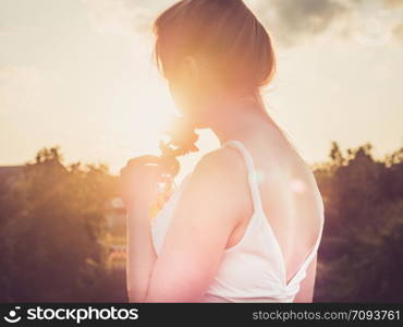 Young mom in a stylish, white dress standing on a background of clear sky and sunlight, holding a beautiful red rose flower in her hands. Concept of a happy family and motherhood. Young mom in a stylish, white dress