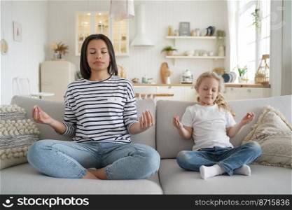 Young mom or babysitter and adopted little daughter practicing yoga together, sitting on sofa at home. Calm mother and kid girl relaxing together in lotus position on couch. Healthy lifestyle concept.. Mom or babysitter and adopted child daughter practicing yoga together at home. Healthy lifestyle
