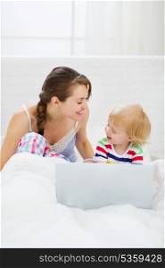Young mother and baby playing with laptop in bed