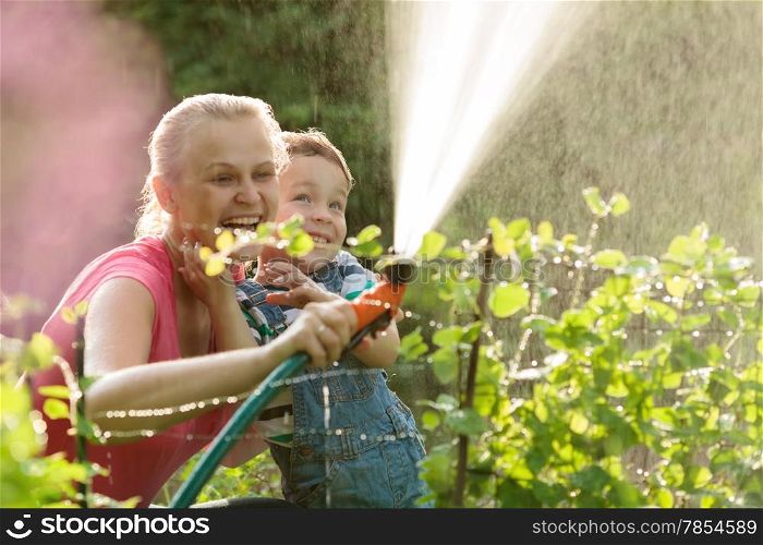 Young mother and cute son playing with water hose in the garden