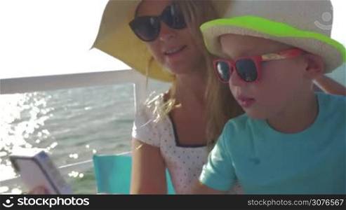 Young mother and her child are doing selfie on the boat. Little boy kisses mom on the cheek, they laugh and look happy on this sunny, summer day
