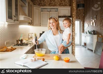 Young mother and her daughter mixing fruits in a bowl. Cute woman and little girl cooking on the kitchen, cake preparation. Happy family prepares sweet dessert, ingredients on the table. Mother and her daughter mixing fruits in a bowl