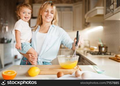 Young mother and her daughter mixing ingredients for cake in a bowl. Cute woman and little girl cooking on the kitchen, pastry preparation. Happy family prepares sweet desserts. Mother and daughter mixing ingredients for cake