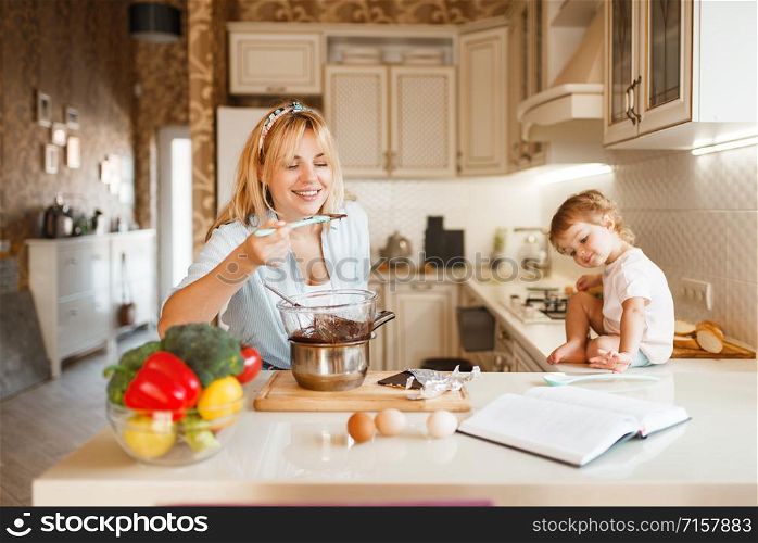 Young mother and her daughter tastes melted chocolate. Cute woman and little girl cooking on the kitchen, pastry preparation. Happy family prepares sweet dessert at the counter. Mother and her daughter tastes melted chocolate
