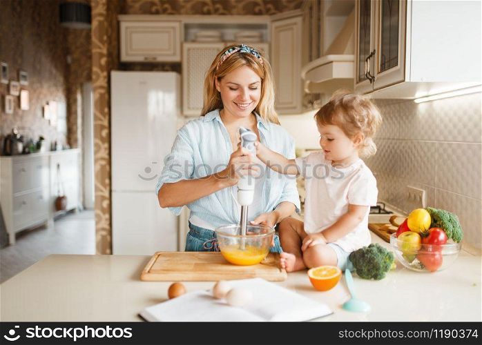 Young mother and her daughter whisk the ingredients for cake with a blender in a bowl. Cute woman and little kid cooking on the kitchen, pastry preparation. Happy family prepares sweets. Mother and daughter whisk ingredients with blender