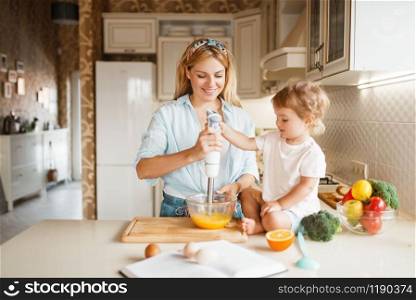 Young mother and her daughter whisk the ingredients for cake with a blender in a bowl. Cute woman and little kid cooking on the kitchen, pastry preparation. Happy family prepares sweets. Mother and daughter whisk ingredients with blender