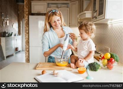 Young mother and her daughter whisk the ingredients for cake with a blender in a bowl. Cute woman and little kid cooking on the kitchen, pastry preparation. Happy family prepares sweet dessert