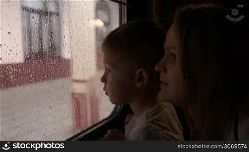 Young mother and her little son in the train looking out the window on a rainy day while passing the station