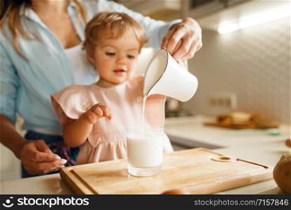 Young mother and kid pours milk into a glass, ingredients for pastry. Woman and little girl cooking on the kitchen, cake preparation. Happy family makes sweet dessert at the counter