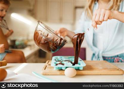 Young mother and kid prepares pastry and tastes melted chocolate. Cute woman and little girl cooking on the kitchen, cake preparation. Happy family makes sweet dessert at the counter. Mother and kid cooking and tastes melted chocolate