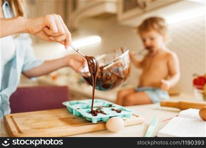 Young mother and kid prepares pastry and tastes melted chocolate. Cute woman and little girl cooking on the kitchen, cake preparation. Happy family makes sweet dessert at the counter