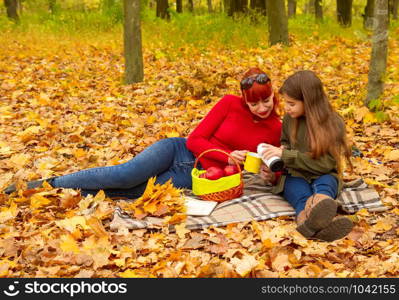 young mother and little daughters in bright sweaters on a picnic in nature drink tea from a thermos. mom and daughter picnic