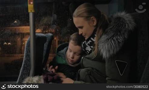 Young mother and son traveling by bus in late winter evening. They passing the time playing on smart phone