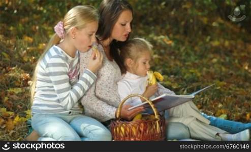 Young mother and two daughters having picnic in the autumn park, reading book