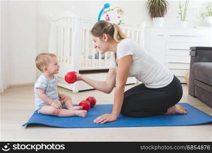 Young mother doing fitness and giving dumbbells to her baby. Concept of family sport