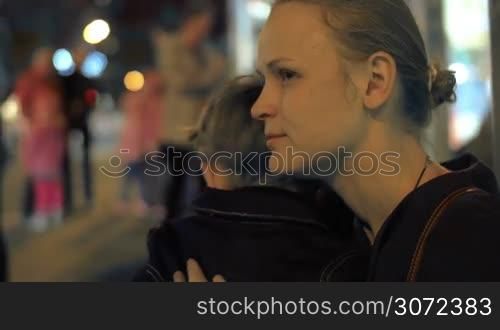 Young mother giving comfort to the little son in the city street in the evening. She embracing him and stroking his back gently