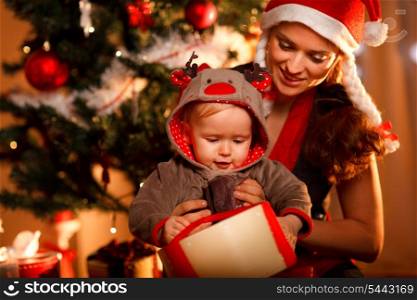 Young mother helping interested baby open present box at Christmas tree&#xA;
