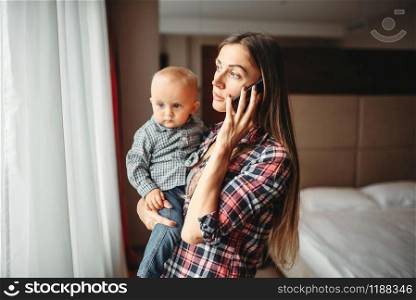 Young mother holding a baby in her arms and talking on the phone indoors. Mom and son happy together at home, togetherness