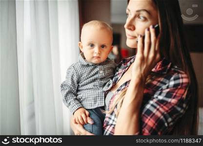 Young mother holding a baby in her arms and talking on the phone indoors. Mom and son happy together at home. Mother holding baby in arms and talking by phone