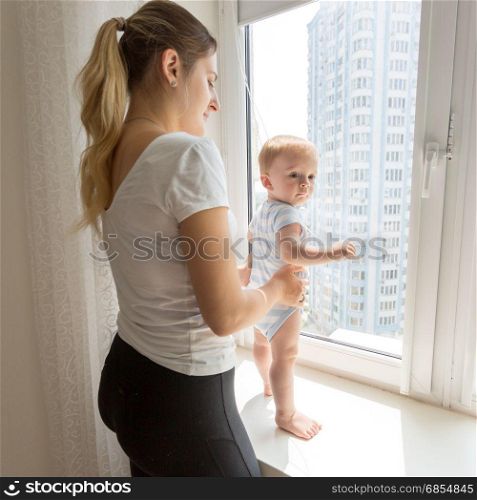 Young mother holding her baby while it is standing on windowsill