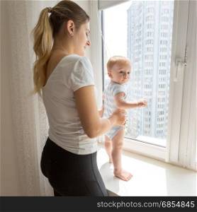 Young mother holding her baby while it is standing on windowsill