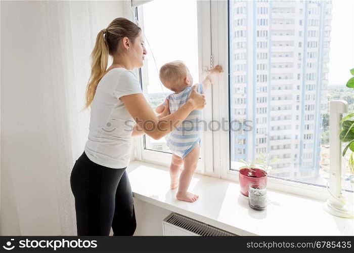 Young mother holding her baby while it is standing on windowsill and looking at window