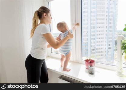 Young mother holding her baby while it is standing on windowsill and looking at window