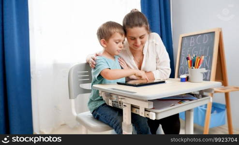 Young mother hugging her little son doing homework and studying on tablet computer at home. CHild using gadget for education and learning.. Young mother hugging her little son doing homework and studying on tablet computer at home. CHild using gadget for education and learning
