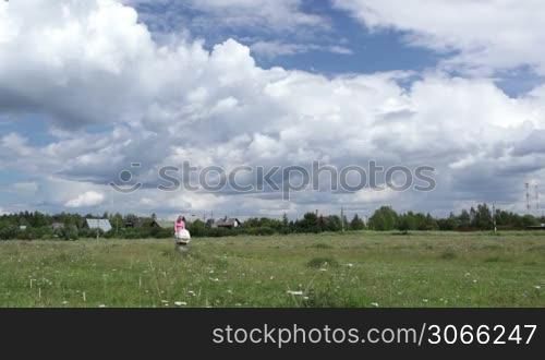 Young mother in a pink T-shirt and shorts with a bag in her hand walking with a buggy across the field on a sunny day. Wide shot. Shot with polarizer.
