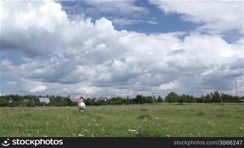 Young mother in a pink T-shirt and shorts with a bag in her hand walking with a buggy across the field on a sunny day. Wide shot. Shot with polarizer.