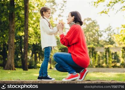 Young mother in warm knitted red sweater plays with her small daughter in park, gives her leaf, enjoy sunny autumn weather. Affectionate mom and little child spend time together outdoor