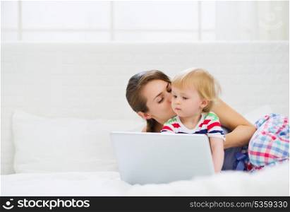 Young mother kissing her modern baby working on laptop