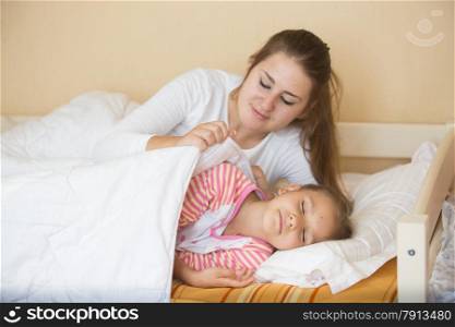 Young mother lying in bed with daughter and covering her with blanket
