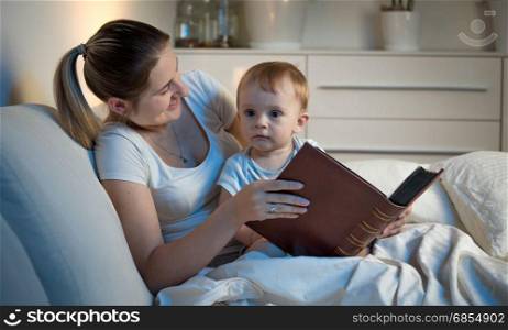 Young mother lying in bed with her baby at night and holding big book
